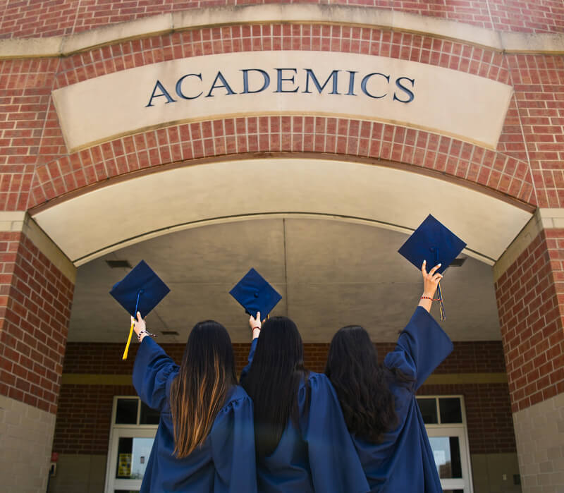 3 girls in graduation gowns facing an academics building.